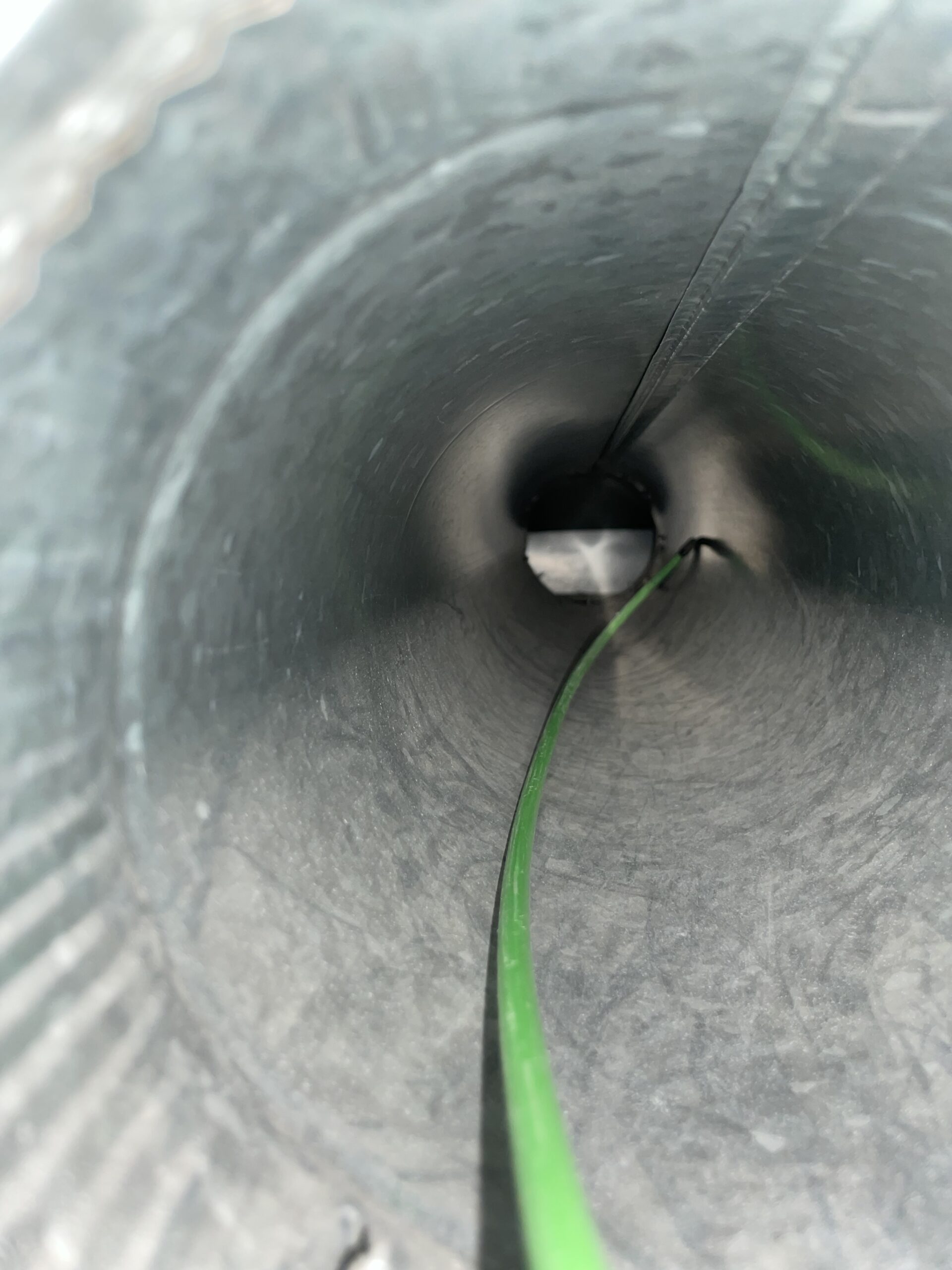 Bright green cleaning brush extends into a dark corrugated air duct with ridged gray walls.