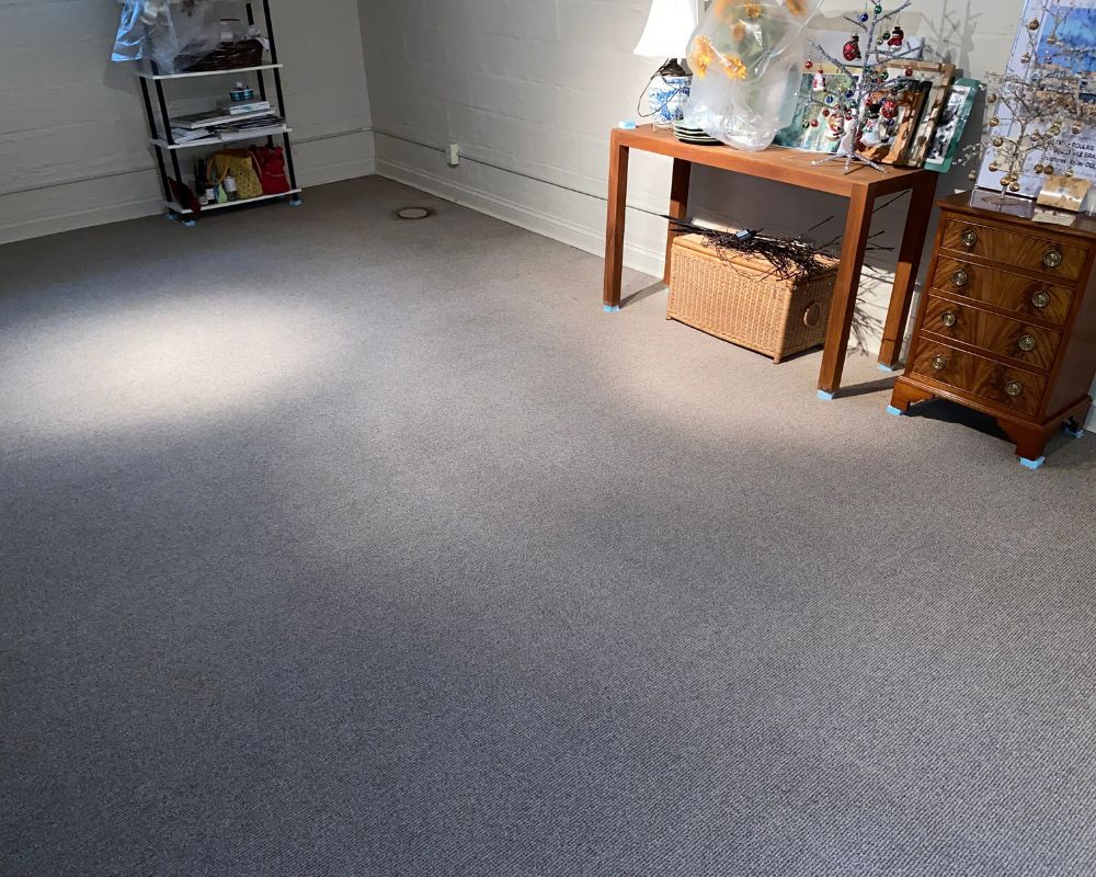 Sunlit gray-carpeted room with wooden table, lamp, chest of drawers, basket, and wall decorations.