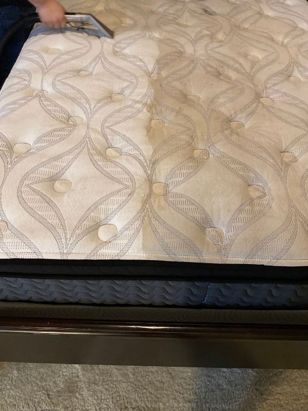 Hand cleaning a patterned mattress with a prominent brown stain above a dark bed frame.