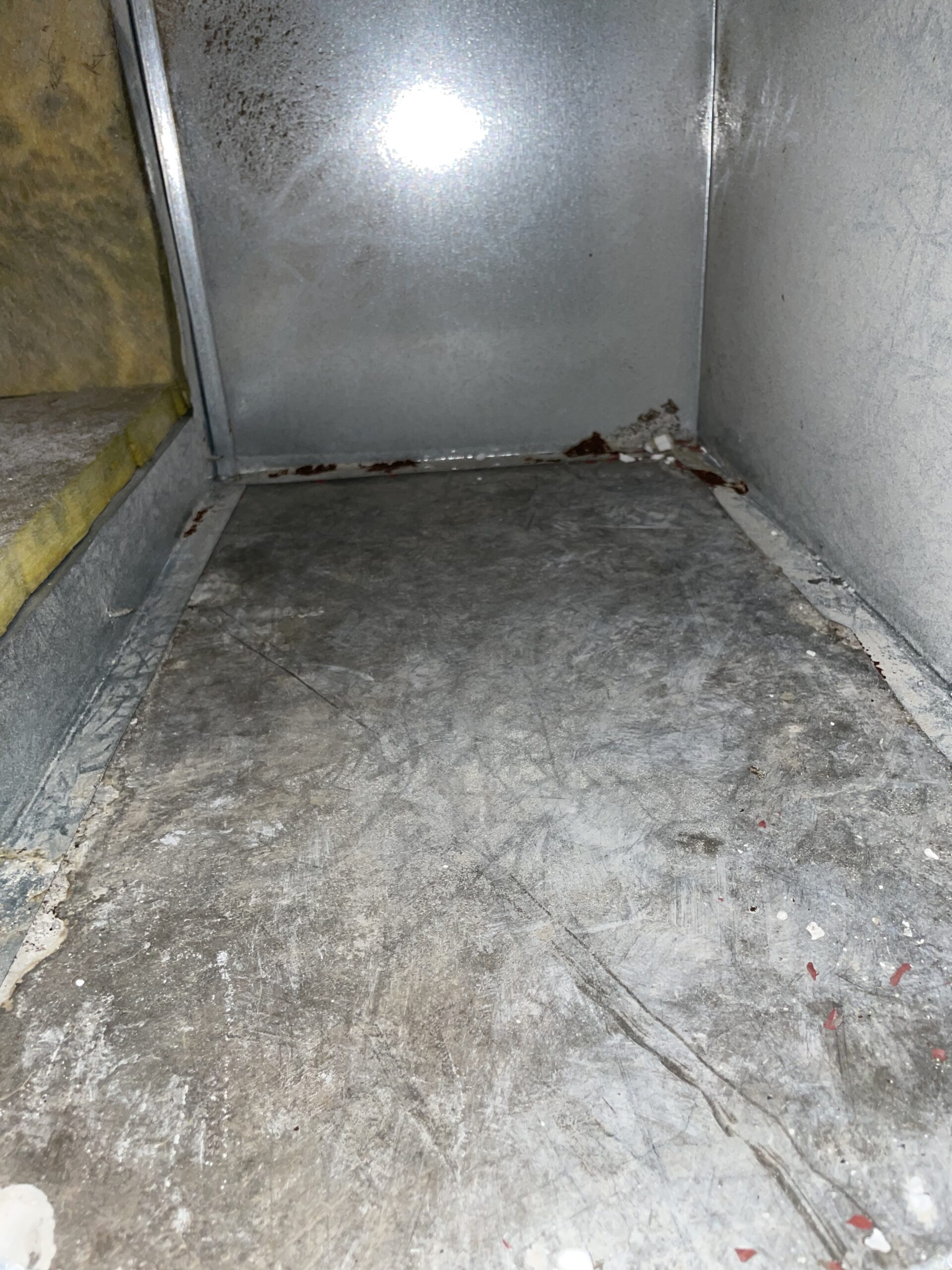 Grimy metal duct interior with stained floor, yellow insulation, scratched panels, and rust buildup.