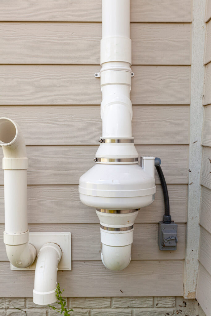 Exterior radon vent pipe assembly with white PVC fittings on beige house siding above foundation.