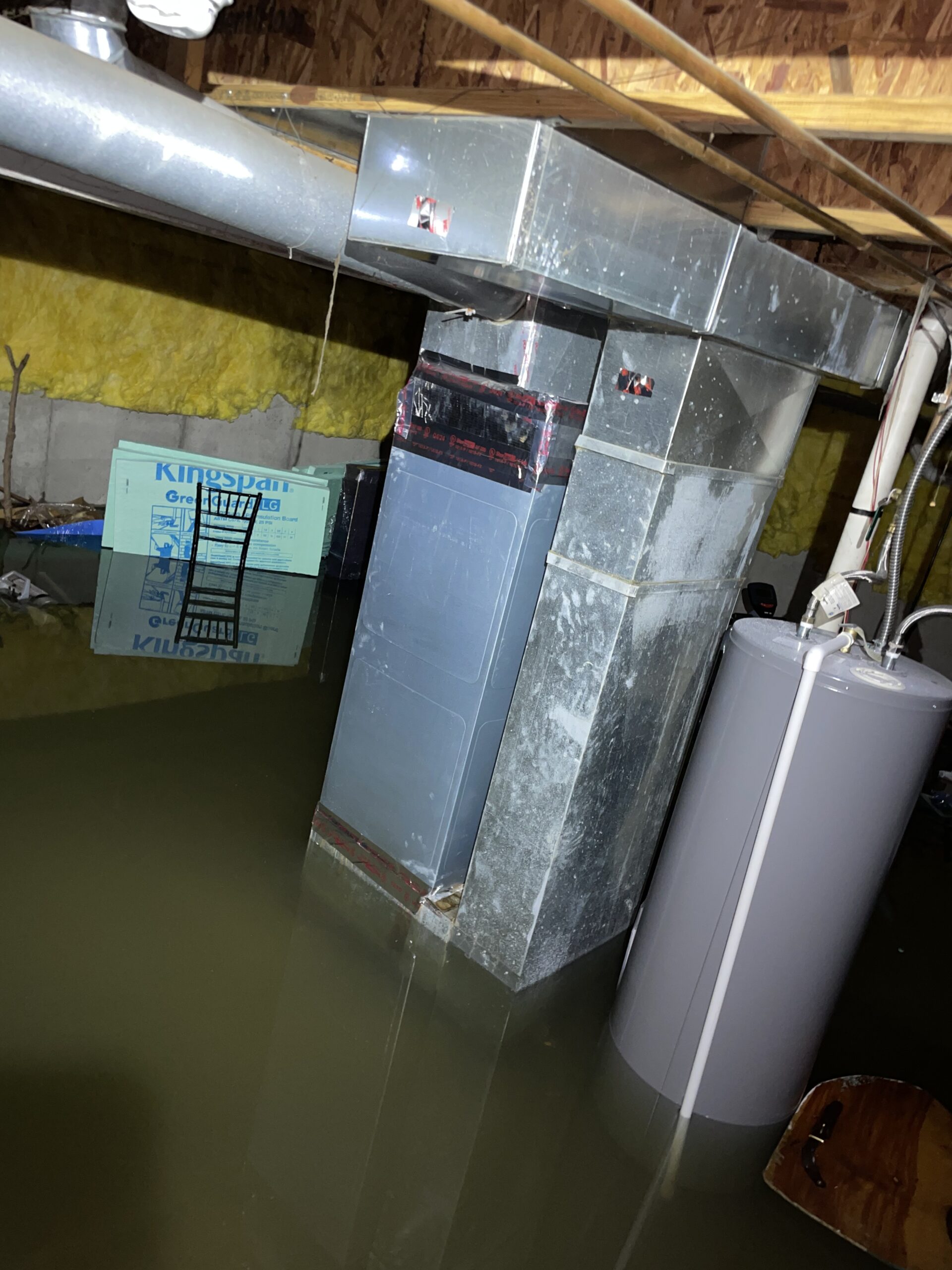 Flooded basement with standing water surrounding a furnace, ductwork, storage tank, and exposed pipes.