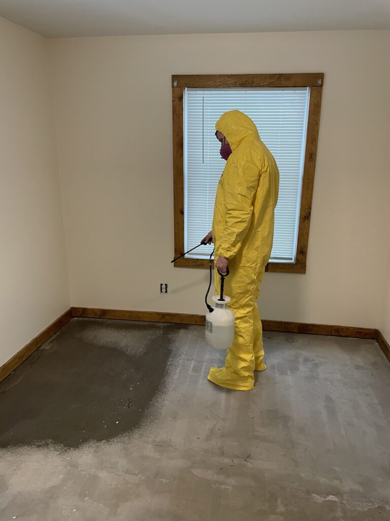 Mold remediation worker in yellow protective suit standing in unfinished room with damaged concrete floor.