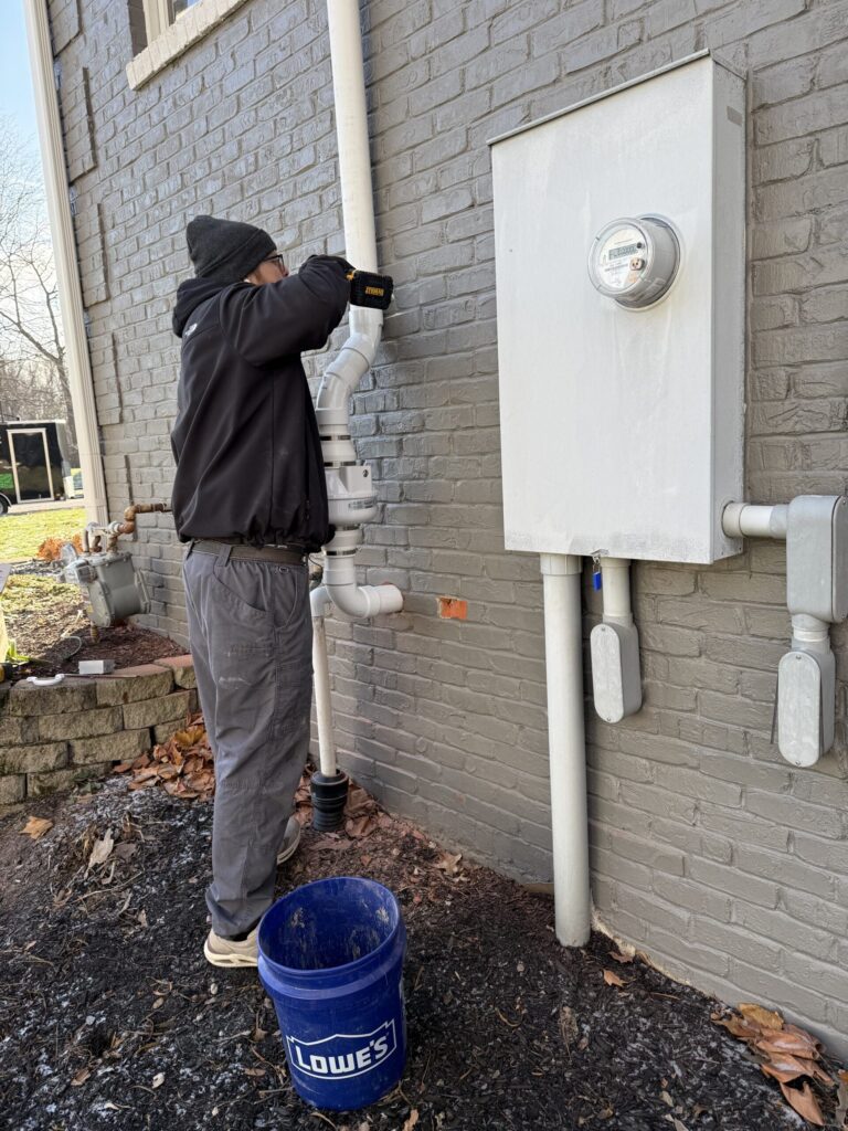 Worker installs a radon mitigation system with white pipes on a gray brick building.