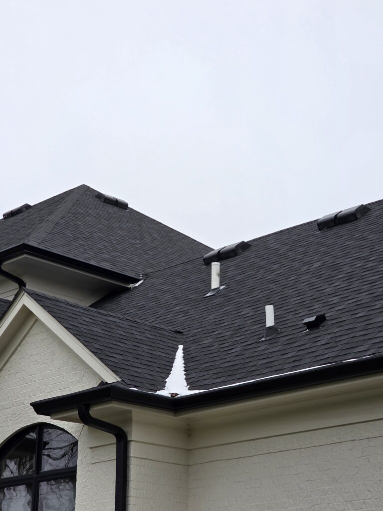 Close-up modern home roof with dark shingles, multiple gables, ridge vents, and white vent pipes.