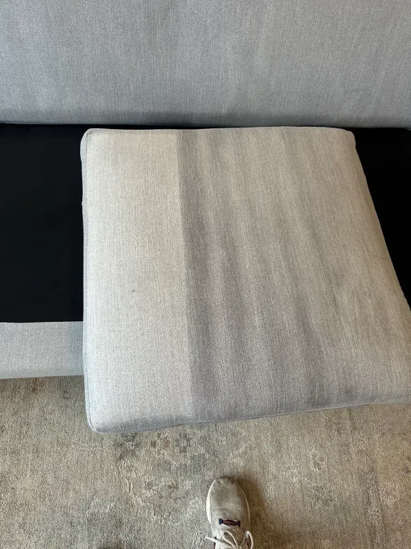 Close-up of a beige sofa cushion during cleaning, showing fabric creases, carpet, and a sneaker.