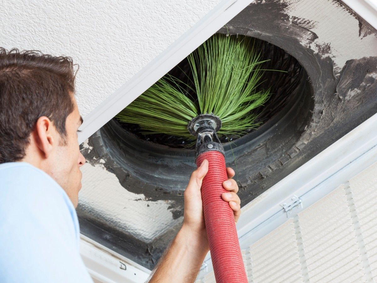 Worker using a rotary brush to clean dust from a large air duct ventilation opening.